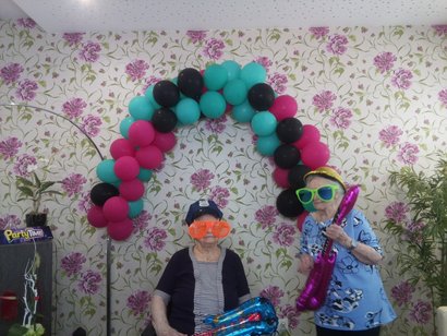 zwei Seniorinnen stehen in Fotobox und halten Luftballons als Musikinstrumente in den Händen.