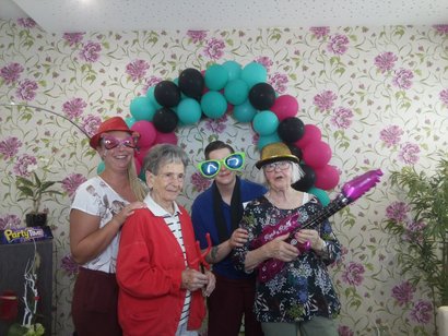 Seniorinnen und ihre Alltagsbegleiterin stehen verkleidet in der Fotobox. Sie tragen große Brillen und halten Luftballons in Form von Instrumenten. Es ist Sommerfest im Seniorenzentrum